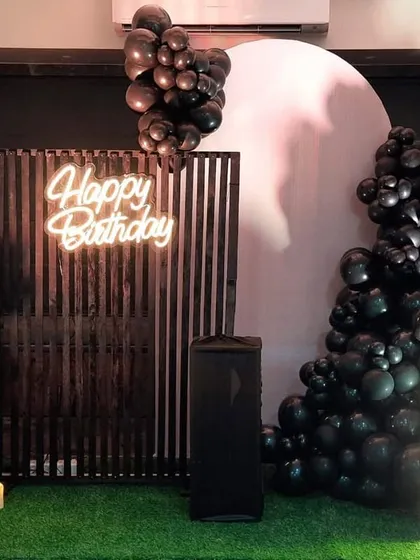 A stylish and modern birthday setup on an artificial grass floor, featuring a black balloon garland, a slatted wooden screen, and a light-up initial.
