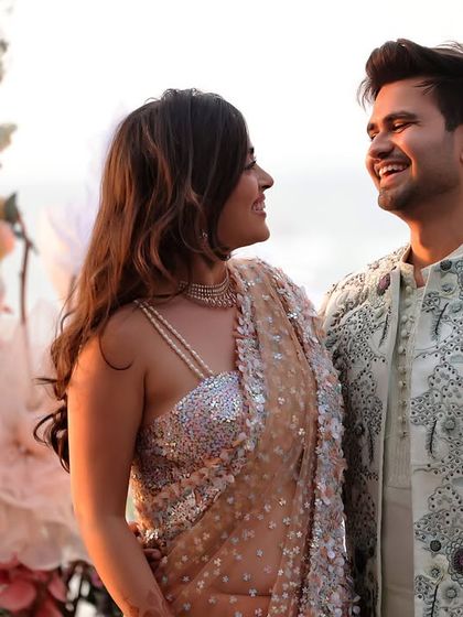 A candid photograph of a couple sharing a laugh during their outdoor engagement shoot. The groom's detailed sherwani and the soft floral background highlight the celebratory feel of the occasion.