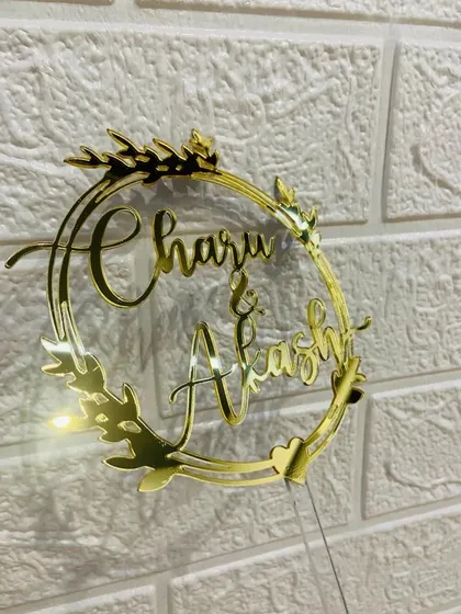 A close-up of the "Charu & Akash" wreath topper against a white brick background.