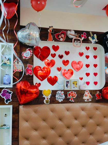 A wall decorated for a romantic surprise, featuring numerous red heart cutouts, heart-shaped foil balloons, and cute couple-themed stickers.