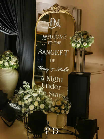 A personalized welcome sign for the 'Night Under the Stars' Sangeet. The elegant mirror with custom calligraphy, flanked by white floral arrangements, sets a sophisticated tone.