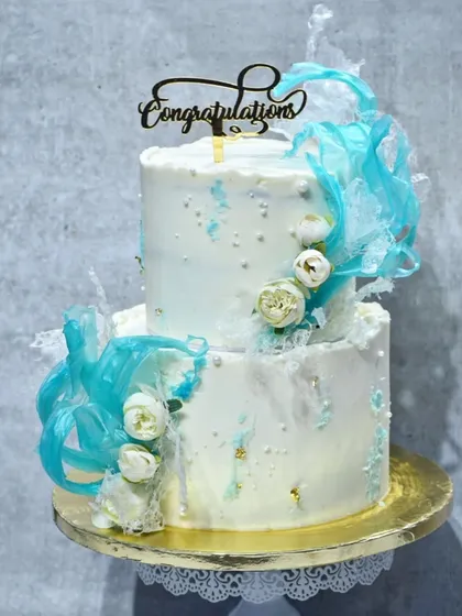 A stunning two-tier cake with a white marble effect, decorated with abstract blue isomalt sails and delicate white flowers. The "Congratulations" topper makes it perfect for celebrating any achievement.