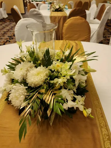 An elegant centerpiece of white chrysanthemums and orchids, accented with metallic gold-painted leaves. The arrangement sits on a golden table runner, creating a cohesive and luxurious look.