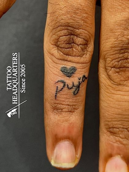A tiny finger tattoo of the name "Puja" with a small heart, a discreet yet meaningful piece.