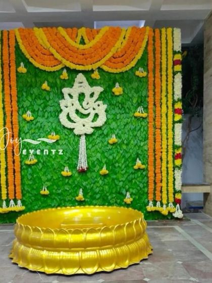 A Ganesha-themed backdrop for a puberty function, crafted with a banana leaf wall and marigold borders. The centerpiece is a Ganesha motif made from white flowers, placed above a golden lotus urli.