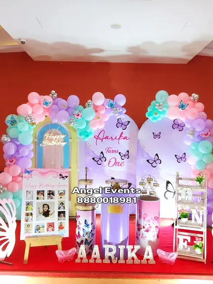 An enchanting butterfly theme in pastel shades of purple, pink, and mint green. The decoration features two arched backdrops, a full balloon garland, custom name lettering, and whimsical butterfly wing props for a magical party atmosphere.