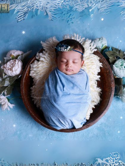 A baby swaddled in blue, nestled in a wooden bowl on a fluffy white blanket, against a wintery blue floral backdrop.
