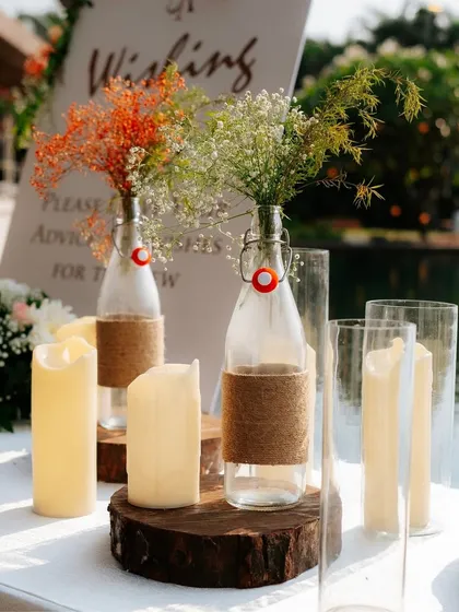 A rustic-chic decor detail featuring wildflowers in cork-wrapped bottles, placed on a wooden slice and surrounded by flameless pillar candles. This is perfect for a relaxed, bohemian-style event.