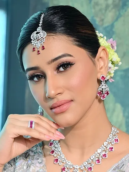 A perfect example of modern bridal glam where less is truly more. This close-up focuses on the flawless, skin-like foundation, soft pink lips, and clean eyeliner. The makeup enhances her features beautifully, complementing the diamond and ruby jewelry for a fresh, elegant reception look.
