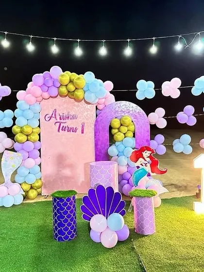 An outdoor Little Mermaid theme for a first birthday. This setup includes a balloon arch, character props, and a marquee number '1' on an artificial grass surface.