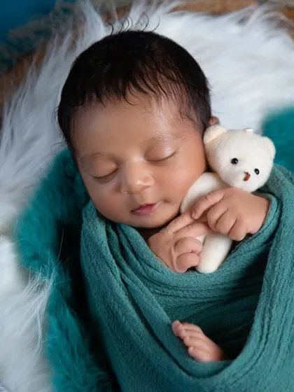A close-up portrait focusing on your baby's delicate features. Here, a newborn is swaddled in a soft teal wrap, peacefully holding a tiny white teddy bear, highlighting the innocence of these early days.