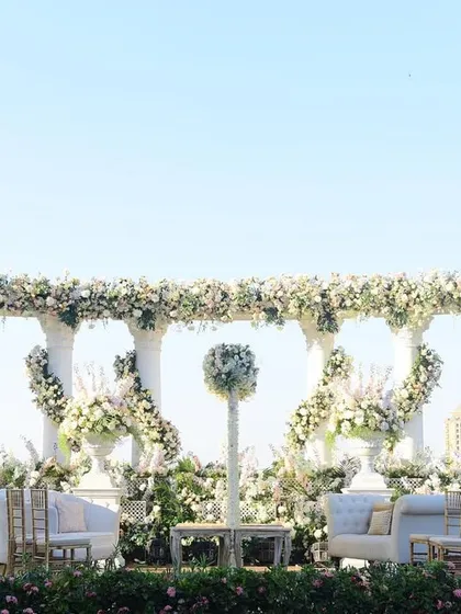 A stunning all-white mandap or stage setup against a clear blue sky. The structure, made of classic white columns, is elegantly wrapped in garlands of white flowers, creating a heavenly look.