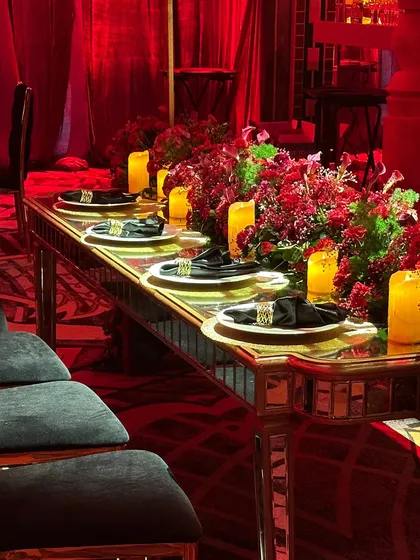 A long, mirrored dining table lined with red florals and candles, reflecting the dramatic red lighting and creating an endless stream of warmth and color.