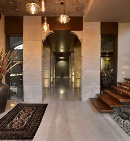 The interior of C Farm features a long corridor defined by stone-clad arches and an exposed concrete ceiling with imprints of palm leaves. A floating wooden staircase adds a modern touch, while a dry riverbed of pebbles below brings a natural element indoors.