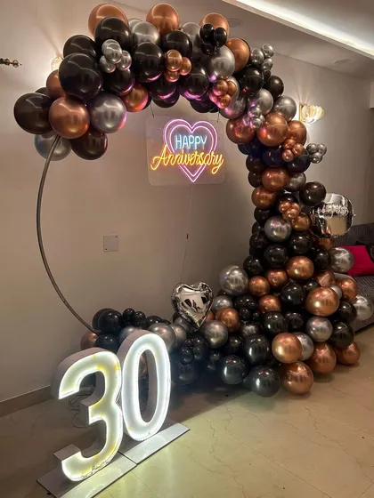 A stylish 30th anniversary party setup. We used a modern color scheme of black, silver, and rose gold for the balloon arrangement on a circular frame, complemented by a 'Happy Anniversary' neon sign and large marquee numbers.