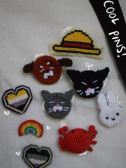 A collection of my cool handmade pins laid out on a tote bag. You can see the variety I offer, from a non-binary pride flag heart and an ace pride flag heart to cute animals like a black cat and a little crab. There's even a straw hat pin for all you anime fans.