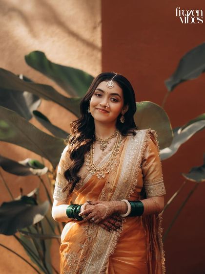 A happy and radiant bride, captured in a moment of quiet joy. The natural background and warm sunlight complement her beautiful golden saree.