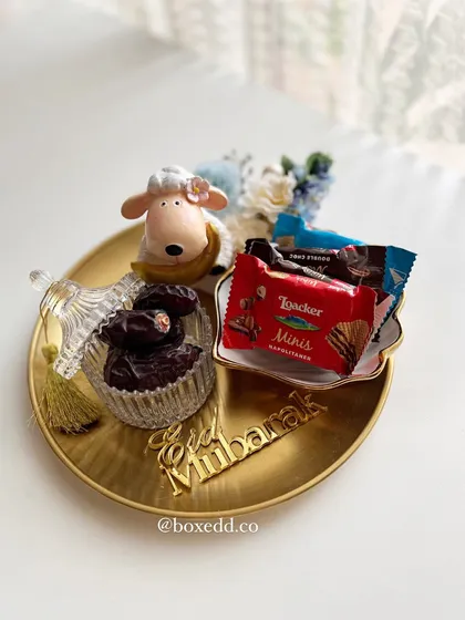 A dreamy Eid-ul-Adha gift platter featuring a cute sheep figurine, a crystal jar of dates, and an acrylic "Eid Mubarak" sign.