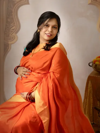 A simple and elegant solo portrait. The mother-to-be is seated, cradling her bump with a warm smile, her vibrant orange saree glowing in the studio light.