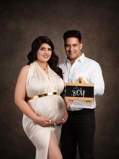 A beautiful gender reveal portrait. The couple stands together, proudly announcing 'It's a Boy' with a small chalkboard sign in this classic, dark-background studio shot.