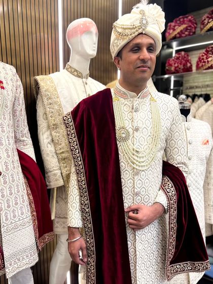 A client trying on a complete groom's ensemble in our store. The look features a white textured sherwani, a pearl mala, and a rich maroon velvet shawl.