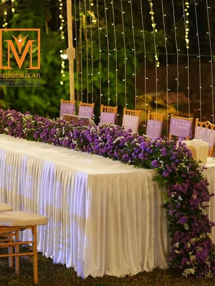 Elegant guest dining for a reception. A long table is decorated with a cascading runner of purple flowers, set under a canopy of fairy lights for a magical, intimate dinner experience.
