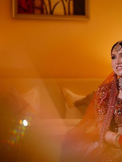 A bride in a maroon lehenga, seated on a bed in a warmly lit hotel room.