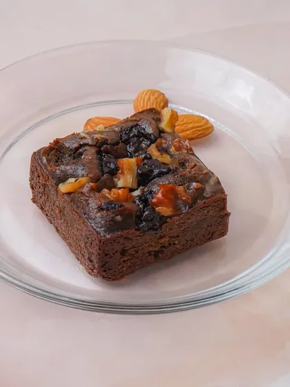 A single square of a dense walnut and almond brownie on a clear glass plate. The simple, clean background puts all the focus on the texture and ingredients of the dessert.