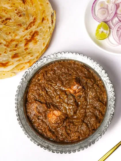 A traditional Mughlai curry served in a decorative metal bowl with a flaky laccha paratha and a side of onions. The top-down flatlay on a white background clearly displays the entire meal.