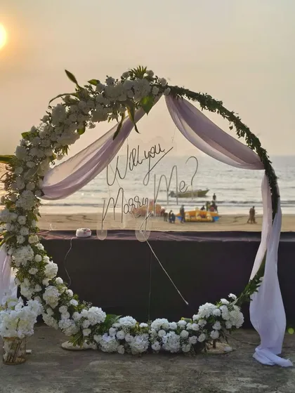 A closer look at the romantic proposal arch. The transparent 'Will you Marry Me?' sign allows the beautiful beach sunset to shine through, while the lush white floral arrangement creates an elegant and timeless frame for the big moment.