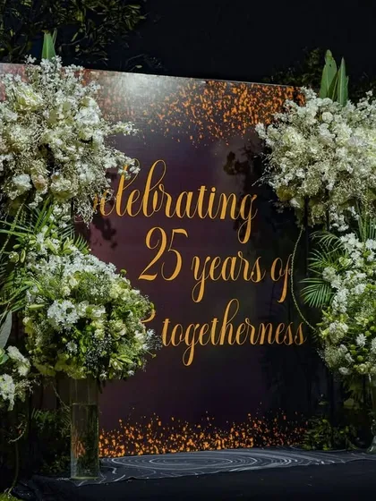 A custom backdrop celebrating "25 years of togetherness," flanked by enormous arrangements of white flowers, creating a perfect photo opportunity for the happy couple and their guests.
