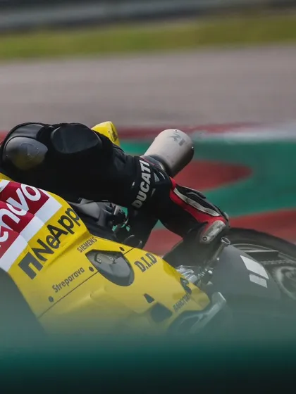 A tight shot of a rider's body and the side of a yellow Ducati, showing the rider's knee puck just above the ground and sponsor logos like Lenovo and NetApp.