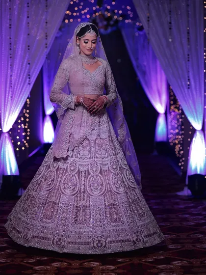 A full-length shot of a bride in a lavender-hued lehenga, looking absolutely regal under the purple stage lighting. Her diamond choker and matha patti complete the monochrome look.