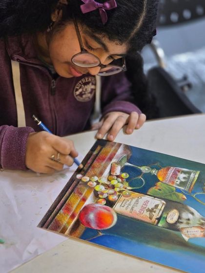 A young artist is deeply focused on her oil pastel drawing, carefully adding details to a still life composition of a wine bottle and fruit.