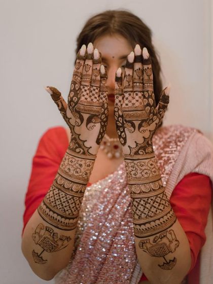 A lovely bride peeking through her hands, which are adorned with intricate bridal mehendi featuring elephants and ceremonial lamps.