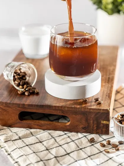 The action of pouring coffee into an iced glass creates a beautiful swirl of color and texture. The rustic wooden tray and scattered beans add to the authentic, cafe-like atmosphere of the shot.