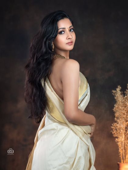 A beautiful back-draped portrait in a traditional white and gold saree. The soft studio lighting and earthy background create a warm, classic feel, perfect for showcasing ethnic wear.