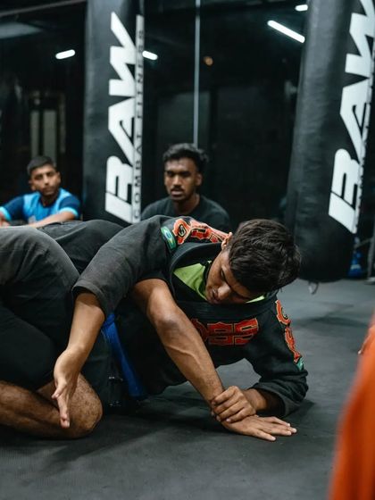 A student in a gi practices a submission hold during a live roll, with other students watching.