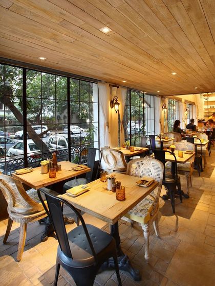 The dining area at Amour Bistro is filled with natural light from large, steel-framed windows. The mix of different chair styles, including classic cane-back and industrial metal chairs, adds to the space's eclectic and charming character.