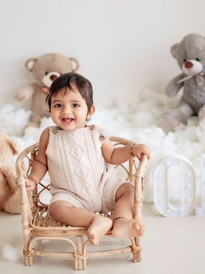 A happy baby boy in a wicker chair, part of a first birthday setup with teddy bears and a "ONE" sign.