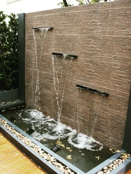 A contemporary wall fountain with three separate spouts. The water flows down a textured stone surface into a basin, creating a multi-sensory experience with both sound and movement.