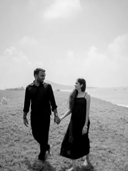 A black and white shot of the couple walking hand-in-hand along a grassy cliff by the sea in Alibag. The composition has a classic, timeless feel.