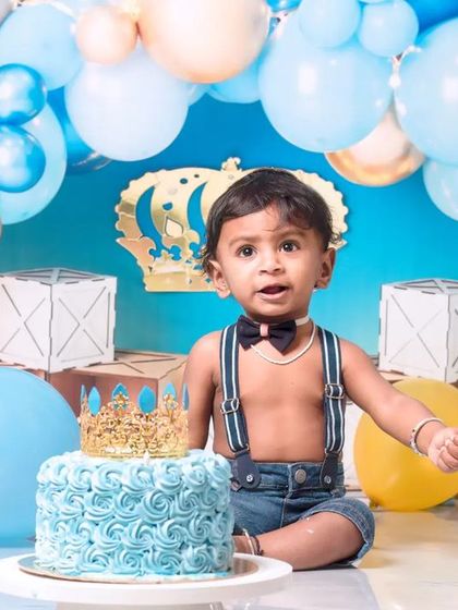 A little prince celebrating his first birthday with a royal blue cake smash. The setup includes a crown, balloons, and a delicious-looking cake ready to be smashed.
