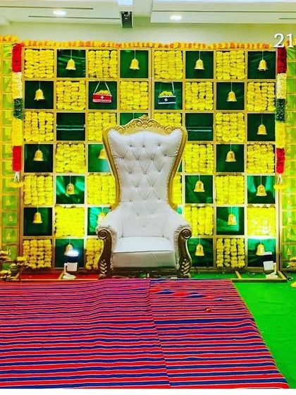 A bright and traditional Seemantham stage with a green and yellow checkerboard backdrop. The decor is accented with hanging bells and a traditional woven mat for the ceremony.