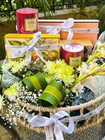 This bespoke hamper was designed to match the bride and groom's green and white wedding theme. It includes a bottle of wine, aromatic candles, and luxury wellness products from Kama Ayurveda, all nestled in a rustic cane basket with fresh white and yellow florals.
