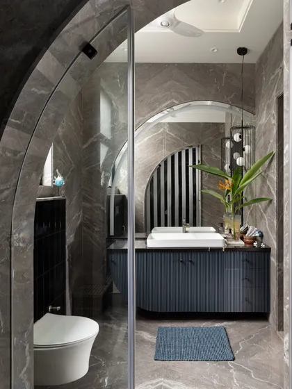 A bold and dramatic bathroom featuring dark grey marble walls in a herringbone pattern. The arched glass shower enclosure and curved vanity soften the strong materials, creating a space that is both moody and elegant.