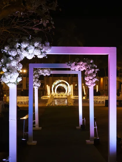 A modern and minimalist entrance gallery for a night function. The series of illuminated square arches adorned with white flowers creates a futuristic and dramatic pathway for guests, leading to the main event area.