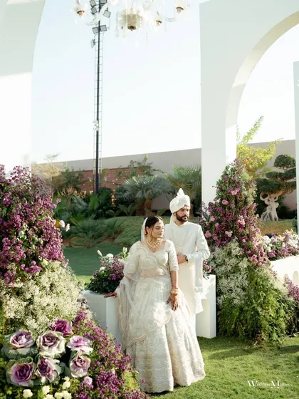 A classic portrait of the couple on their wedding day, standing amidst lush floral decor. Their coordinated white outfits and serene expressions create a timeless and elegant image.