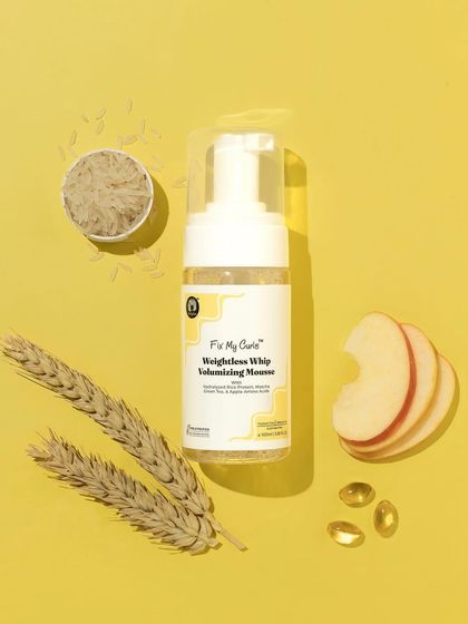 A flatlay composition that showcases the natural ingredients of a volumizing mousse. The arrangement of wheat, rice, and apple slices on a yellow background feels wholesome and effective.
