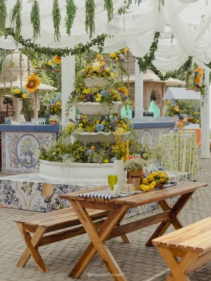 A wide shot of the Mehendi setup, with picnic tables, a floral fountain, and bar counters with custom tile prints, all under a canopy of white drapes and hanging greenery.